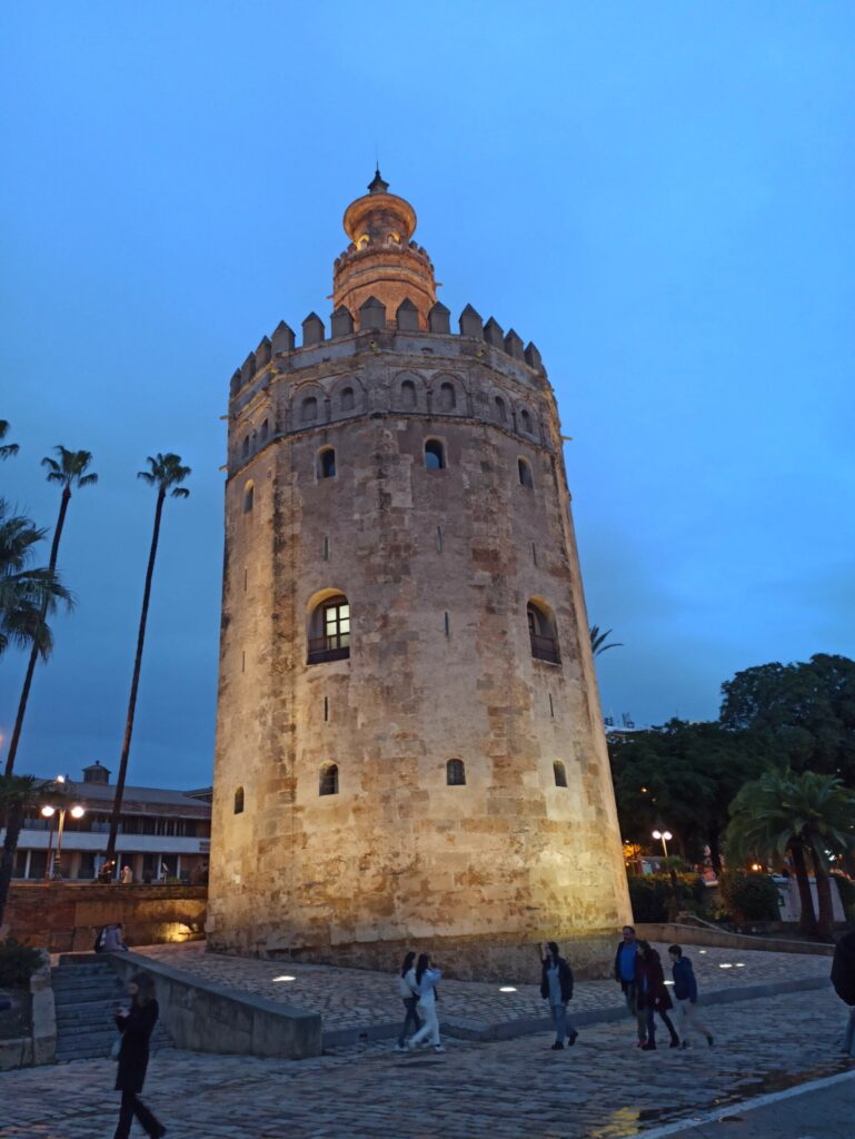 Torre del Oro de Sevilla iluminada al atardecer, con palmeras y gente paseando